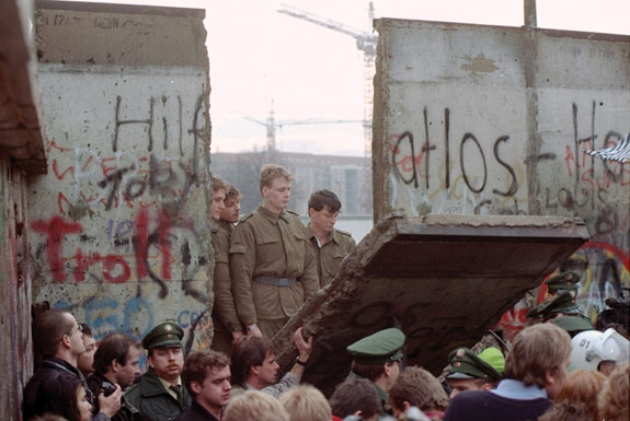 East German border guards look through a hole in the Berlin Wall after demonstrators pulled down a segment of it at the Brandenburg gate, 11 November 1989.